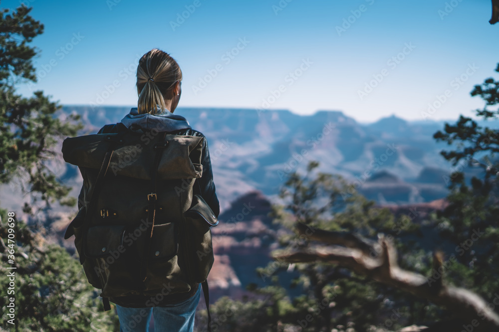 Back view of blonde female explorer with backpack standing in forest enjoying beautiful scenery of mountains scape on active weekend getaway, woman traveler explorer looking at hills on journey