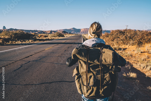 Back view of female traveler passing wild lands near asphalt road exploring southwest of USA,hipster girl enjoying active lifestyle with backpack walking on roadway having journey with hitchhiking