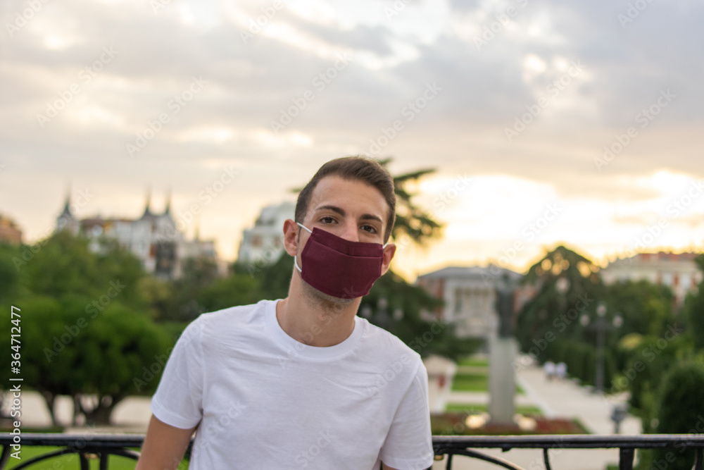 Photo of a young and attractive man with basic and modern clothes in summer wearing a reusable face mask and standing in the park Photo of a young and attractive man with basic and modern clothes in summer wearing a reusable face mask and standing in the park