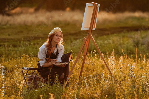  a girl artist with long red hair draws on an easel with a brush against the background of the sunset