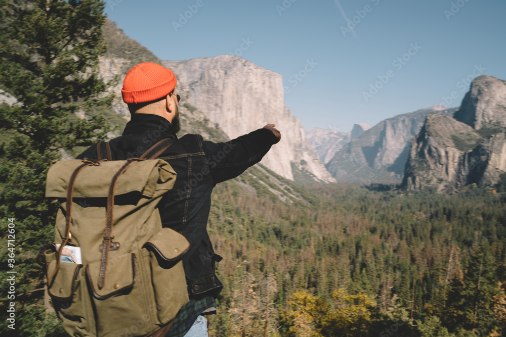 Male traveler with rucksack pointing on high mountain pear looking at beautiful nature and forests in Yosemite national park,back view of hipster guy hiking during tour and active summer vacation. Male traveler with rucksack pointing on high mountain pear looking at beautiful nature and forests in Yosemite national park,back view of hipster guy hiking during tour and active summer vacation.