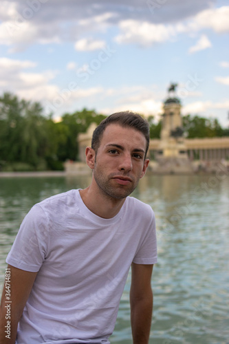 Photo of a young and attractive man with a white t/shirt standing next to the lagoon in Retiro park, Madrid