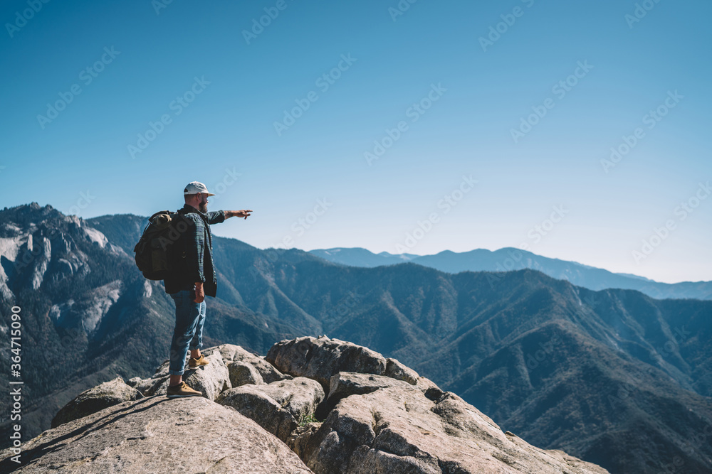Young male traveler with rucksack pointing on mountain peaks standing on rock cliff during hiking tour, guy wanderlust exploring national park landscape showing on location distance from hill.
