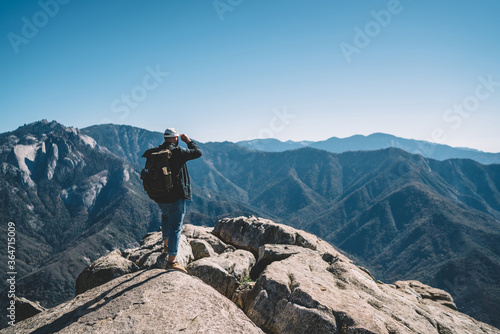 Back view of hipster guy wanderlust enjoying breathtaking scenery from high rock hill, male traveler with backpack recreating explore wild nature looking at mountain peaks landscape 