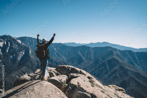 Back view of male traveler celebrating achievement of getting on mountain top destination standing with raised hands,freedom concept of amazed guy wanderlust excited with breathtaking natural scenery.