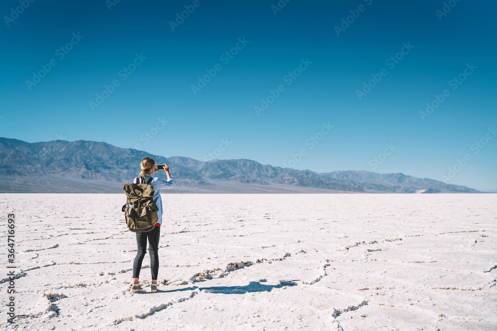 Female traveler with backpack making photo on smartphone of beautiful scenery of arid landscape of dry lake in USA, woman exploring lowest point of North America taking picture of unique environment