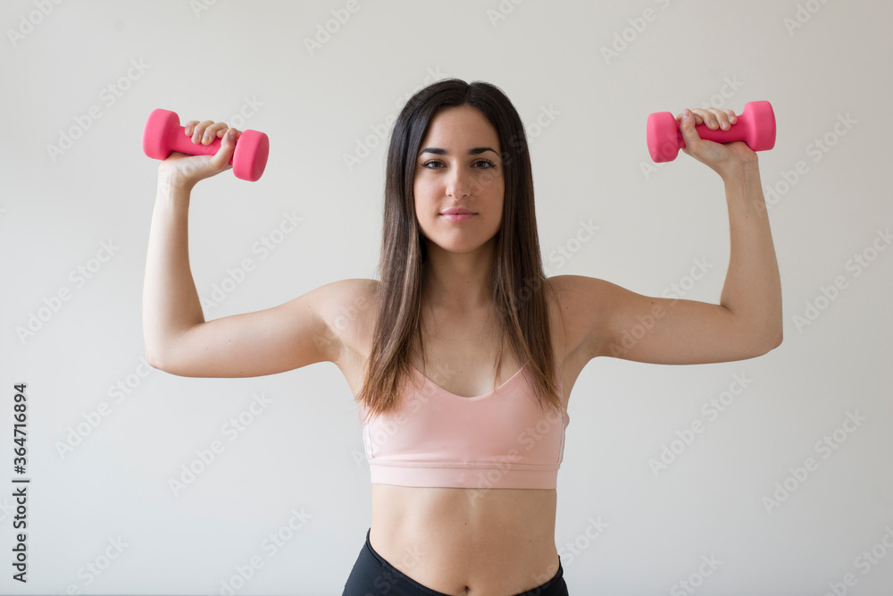Young brunette woman exercising with pink dumbbells in front of a white background Young brunette woman exercising with pink dumbbells in front of a white background
