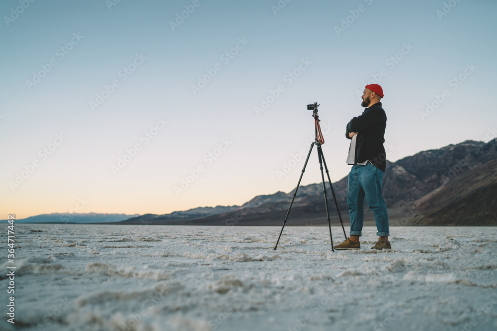 Confident hipster guy with standing with crossed arms satisfied with modern equipment for shooting breathtaking landscape scenery in desert, male photographer using tripod during  expedition