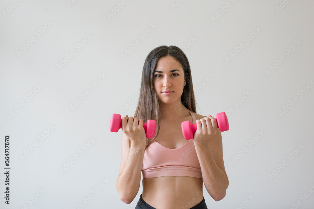 Young brunette woman exercising with pink dumbbells in front of a white background Young brunette woman exercising with pink dumbbells in front of a white background