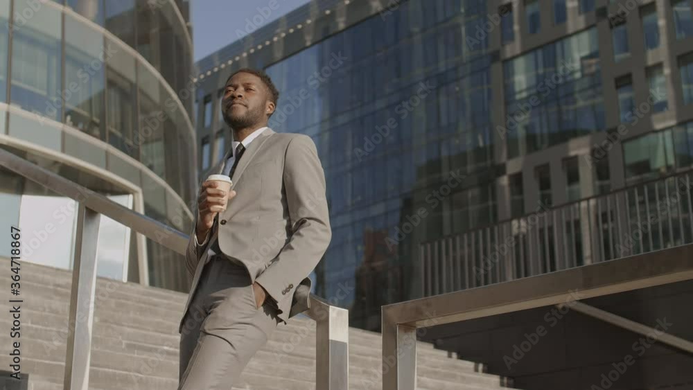 Medium shot of good-looking African businessman wearing elegant clothes standing outdoors on stairs leaning on railing, drinking coffee and looking around Medium shot of good-looking African businessman wearing elegant clothes standing outdoors on stairs leaning on railing, drinking coffee and looking around