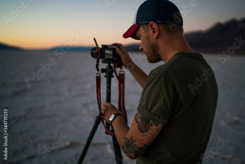 Fototapeta Naklejka Na Ścianę i Meble -  Skilled caucasian male photographer standing near camera and tripod shooting video of beautiful sunset in desert on horizon,hipster guy making photos in dusk in journey to death valley with equipment.