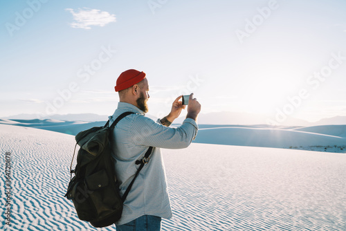 Trandy dressed hipster guy tourist making photo of beautiful sunset and landscape of bunes during hiking tour on vacations, male wanderlust influencer shooting video for sharing in travel blog photo