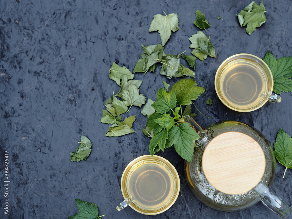 Herbal summer drug background.Tea in a glass teapot and cups with black currant leaves on a black wooden background. Herbal summer drug background.Tea in a glass teapot and cups with black currant leaves on a black wooden background.