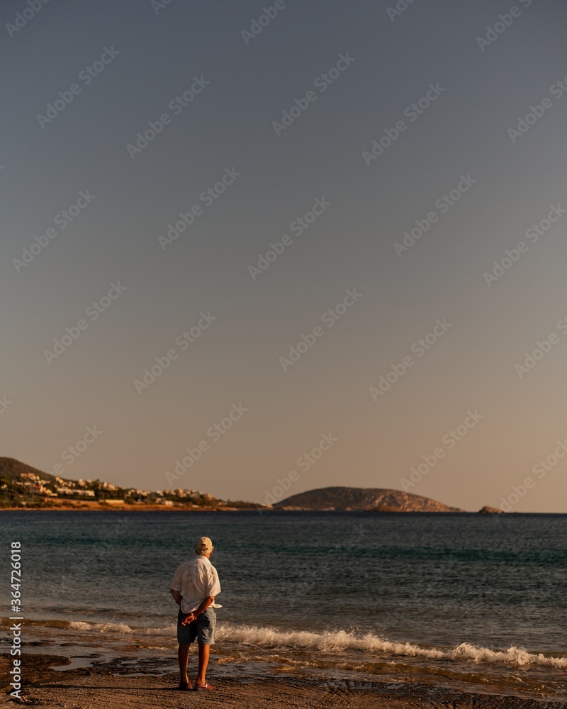 Vertical shot of a male standing at the beach admiring the beauty of the sea under the clear sky