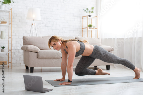 Fitness workout of sporty woman. Exercises for legs on mat with laptop in living room interior