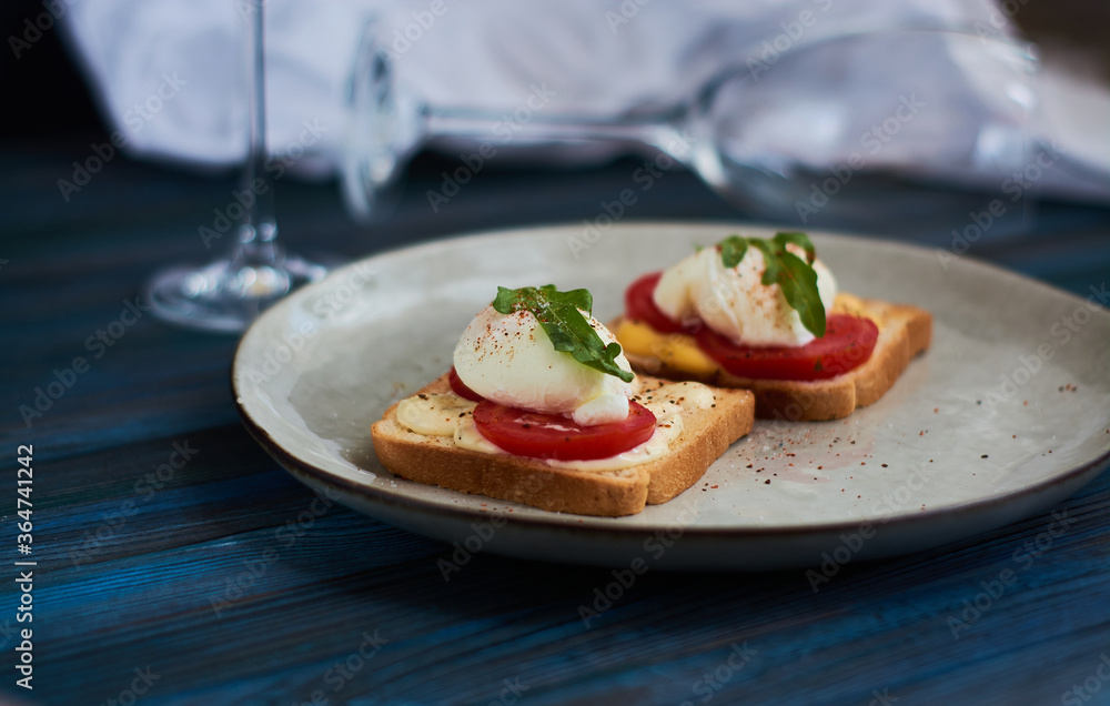Large gray ceramic plate with toast and poached eggs, greens, tomatoes, sauce. Tasty breakfast. Glasses of wine. Large gray ceramic plate with toast and poached eggs, greens, tomatoes, sauce. Tasty breakfast. Glasses of wine.