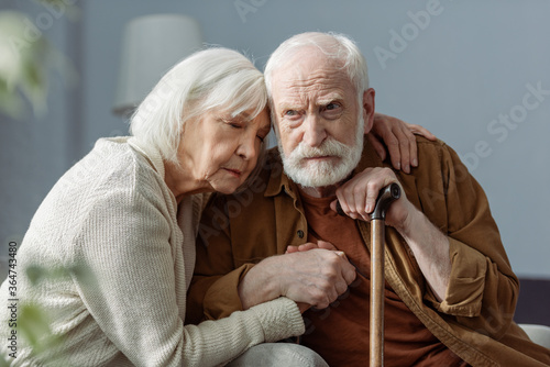 senior woman with closed eyes hugging husband, sick on dementia, and holding his hand photo