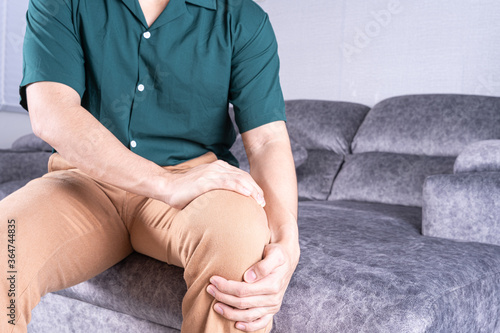 Young man suffering from knee pain while sitting on sofa at home. Healthcare medical or daily life concept.