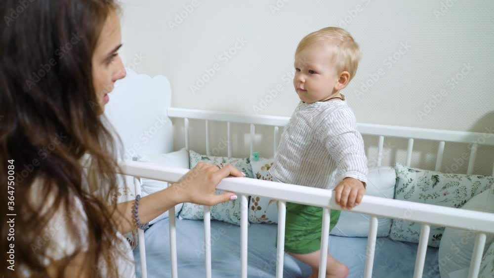 Baby boy standing in crib and happy mother watching him Baby boy standing in crib and happy mother watching him