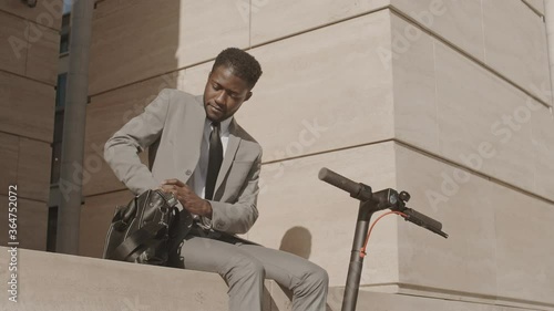Medium shot of young African man sitting in city center near his electric scooter, taking cellphone out of bag and answering call