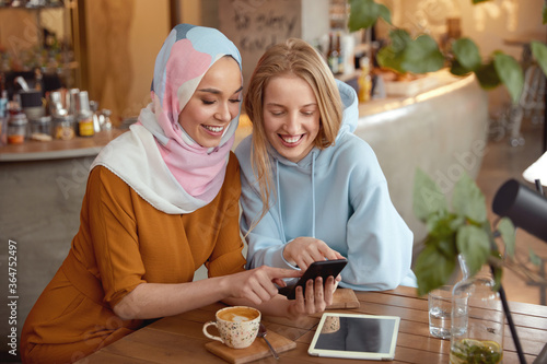Friends Meeting At Coffee Shop. Young Girls Chatting In Cafe. Smiling Women Looking At Smartphone Screen. Different Ethnicity Female Spending Time In Bistro. 