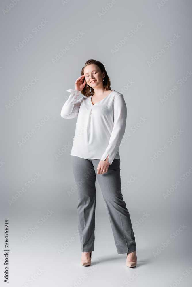 full length view of stylish overweight girl touching hair while posing with closed eyes on grey full length view of stylish overweight girl touching hair while posing with closed eyes on grey