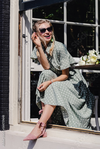 Canvas Print Close-up portrait of laughing blonde girl in pale green long dress with sunglasses, nice hair and beautiful shoes with ankle ties looking out of window of the city cafe