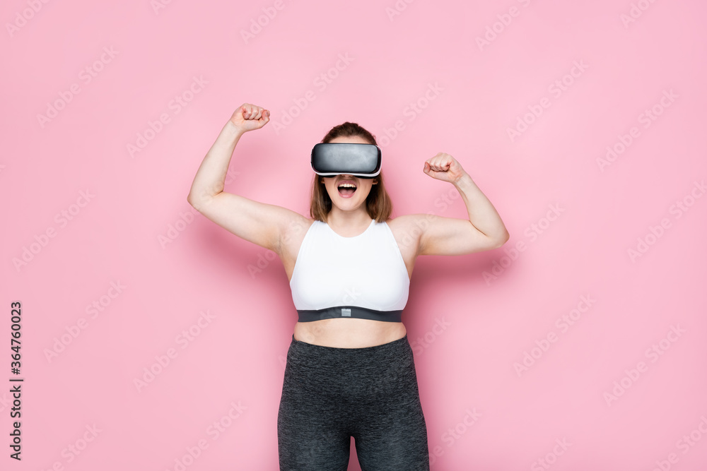 excited size plus girl in vr headset and sportswear showing winner gesture on pink excited size plus girl in vr headset and sportswear showing winner gesture on pink