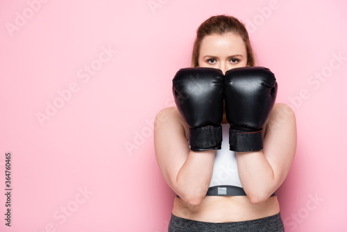 confident overweight girl obscuring face with boxing gloves while looking at camera on pink