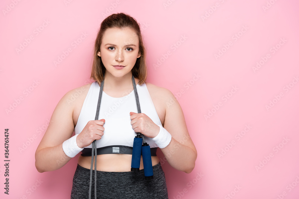 confident overweight girl with jumping rope around neck smiling at camera on pink