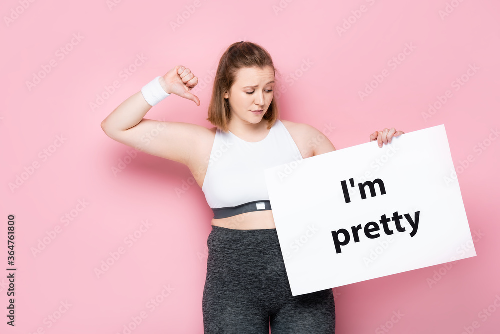 offended overweight girl holding placard with I am pretty inscription and showing thumb down on pink offended overweight girl holding placard with I am pretty inscription and showing thumb down on pink
