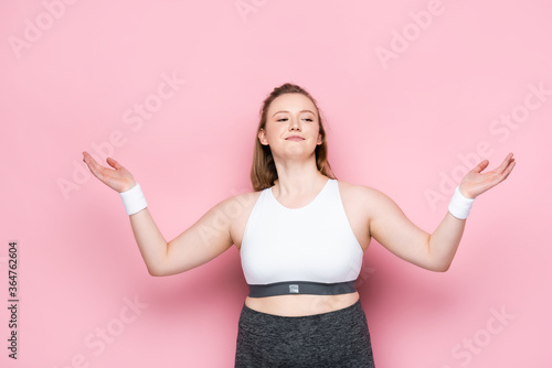 smiling overweight girl in sportswear standing with open arms on pink