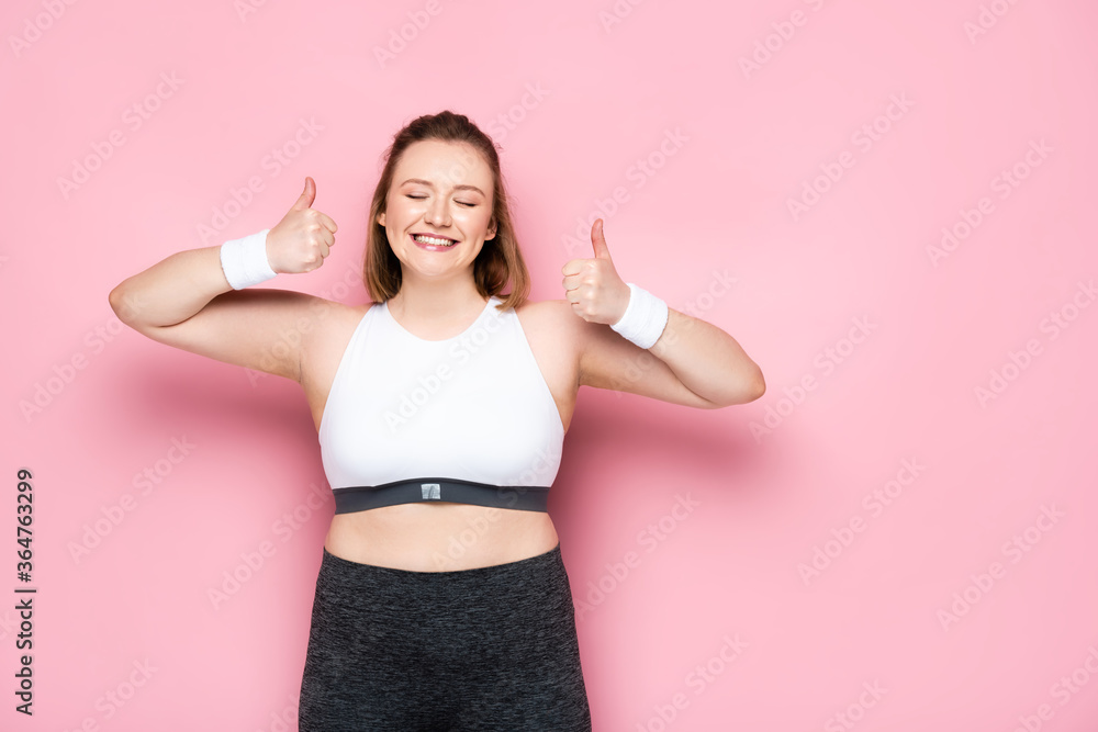 excited overweight girl with closed eyes showing thumbs up on pink
