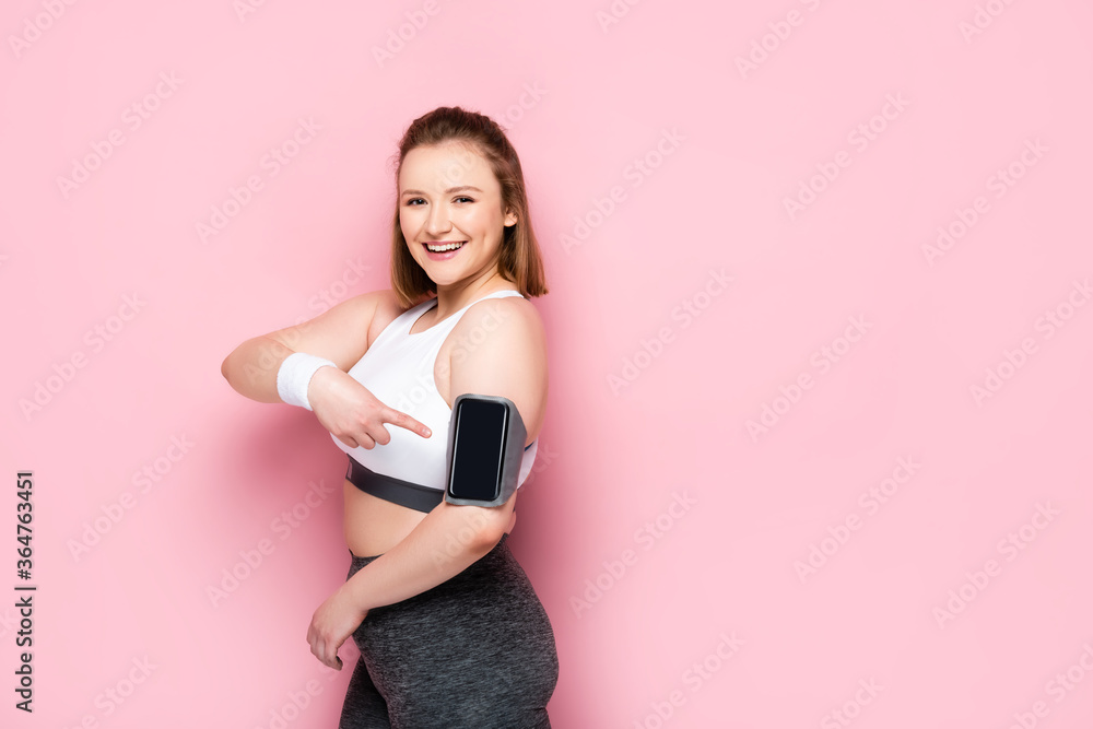 happy overweight girl pointing with finger at smartphone in armband on pink happy overweight girl pointing with finger at smartphone in armband on pink