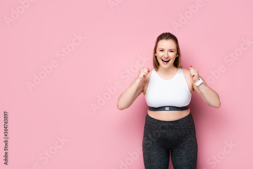 excited overweight girl in sportswear showing winner gesture on pink