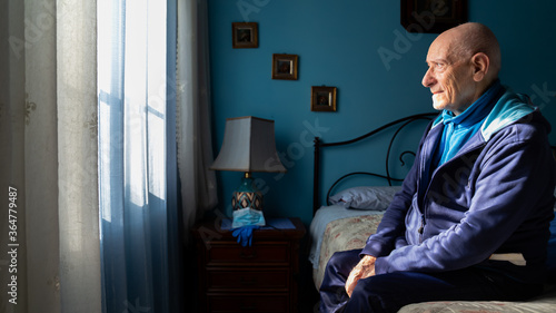 Covid-19 Profile view of an old man sitting on his bed looking sad at the window