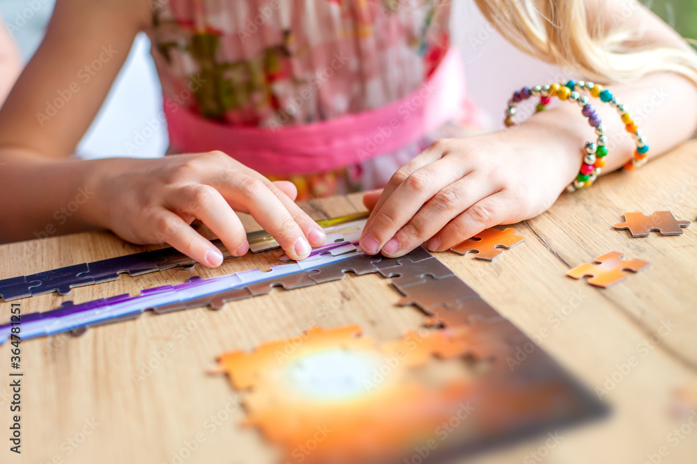 Fototapeta premium A girl sits at a table and collects a puzzle at home. Hands close up. Learning through the game. Learning the concentration of children. We play at home with parents during bad weather and quarantine