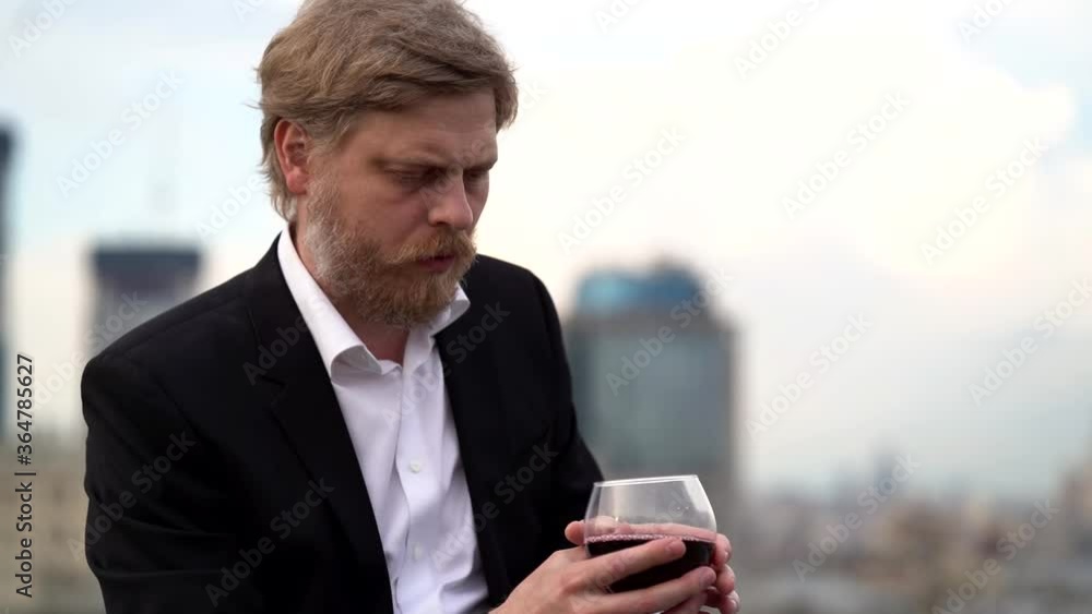 bearded sad man is drinking wine alone on roof in city, abandoned and desperate