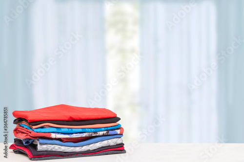 Stack colorful clothes. Pile of folded cotton t-shirts or shirts on a bright table with space for your display product montage over blurred curtains background. Summer fashion.