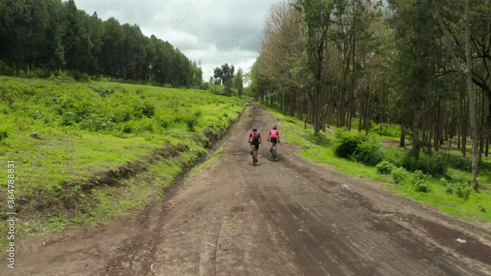 Aerial drone shot flying above marathon athletes on mountainbikes cycling on a dirt road through grass field and farmland very heavy workout in jungle forest Tanzania following shot 4k Aerial drone shot flying above marathon athletes on mountainbikes cycling on a dirt road through grass field and farmland very heavy workout in jungle forest Tanzania following shot 4k