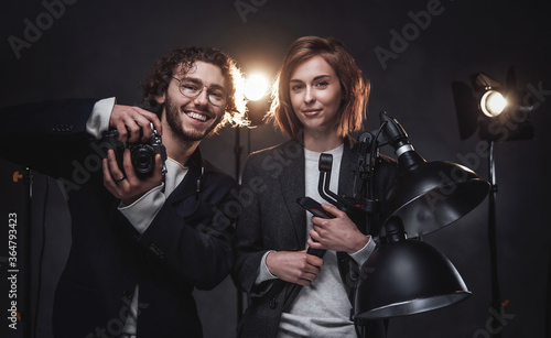 Happy professional photographers in dark studio. Handsome man and beautiful young woman holds a digital camera and lightning equipment