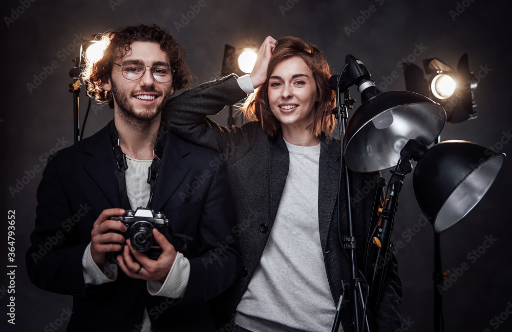 Happy professional photographers in dark studio. Handsome man and beautiful young woman holds a digital camera and lightning equipment