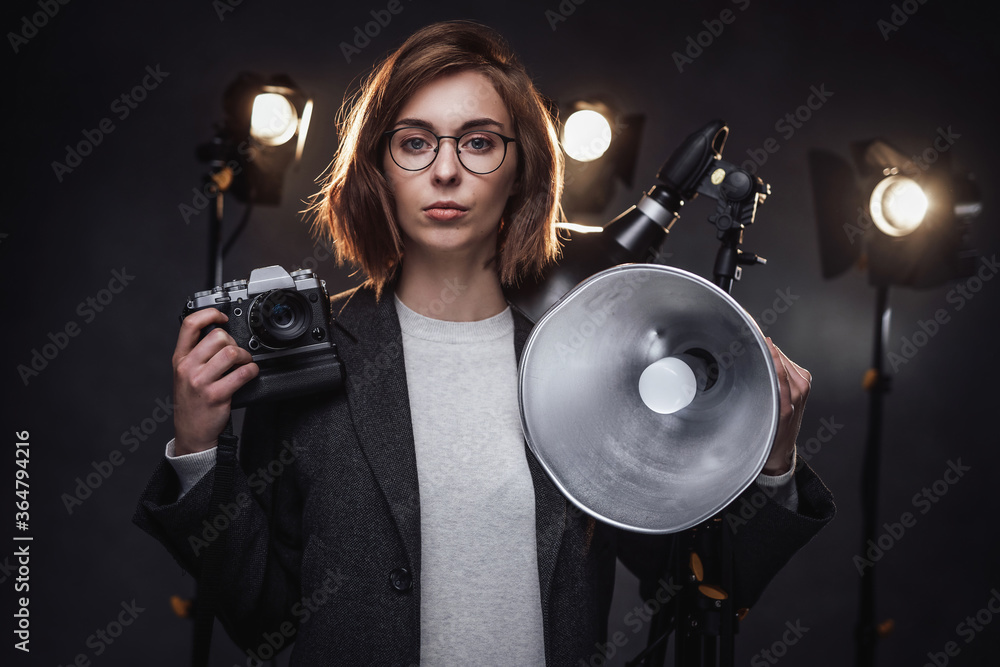 Fototapeta premium Portrait of a beautiful redhead female photographer holds a digital camera and looking on the camera. Studio shot on dark background with lighting equipment