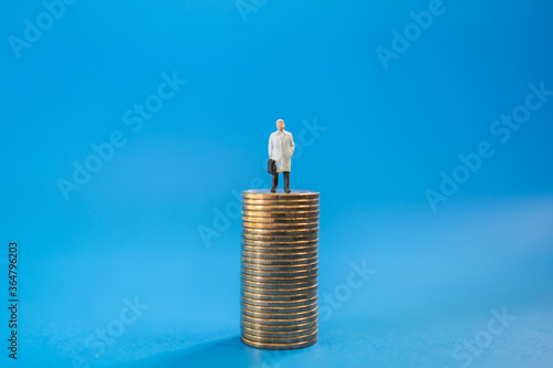 Business, Money Investment and Planning Concept. Close up of businessman miniature people figure with handbag standing on top of stack of gold coins on blue background with copy sapce.