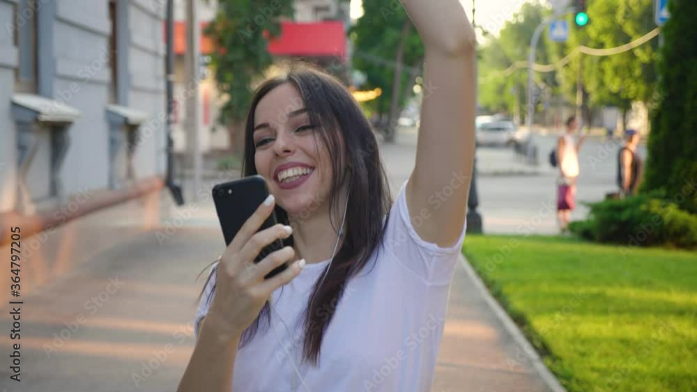 Pretty young woman enjoying her favorite music, dancing and walking down the street. Beautiful young female holding mobile phone listening to music by earphone at city urban background. Steadicam shot Pretty young woman enjoying her favorite music, dancing and walking down the street. Beautiful young female holding mobile phone listening to music by earphone at city urban background. Steadicam shot