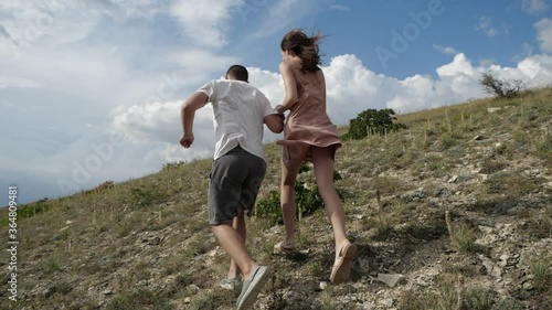 A young couple holding hands runs up a rocky slope to meet lush clouds. A guy in a white T-shirt and a girl in a pink dress with her hair flying in the wind run down the hill
