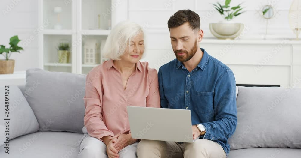Caucasian young handsome adult son teaching his old gray-haired mother to use laptop on couch in cozy living room. Senior woman using computer and asking questions when man explaining and typing. Caucasian young handsome adult son teaching his old gray-haired mother to use laptop on couch in cozy living room. Senior woman using computer and asking questions when man explaining and typing.