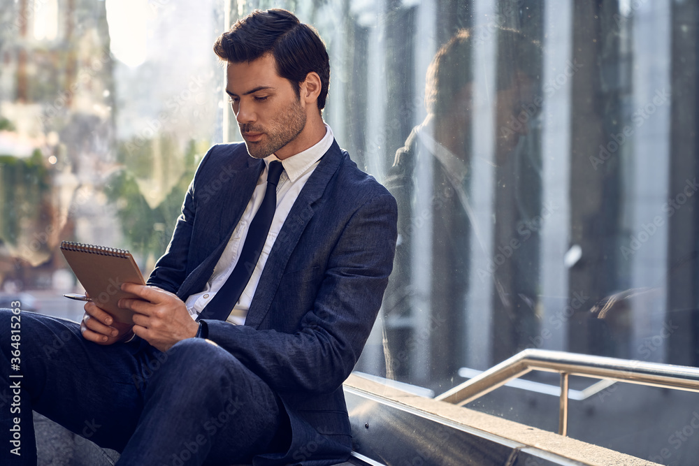Confident happy young entrepreneur in formal suit writing in notebook while sitting at benche outdoors and looking away Confident happy young entrepreneur in formal suit writing in notebook while sitting at benche outdoors and looking away