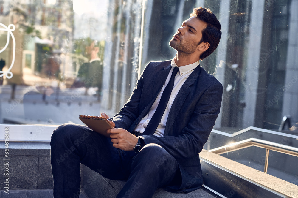 Confident happy young entrepreneur in formal suit writing in notebook while sitting at benche outdoors and looking away Confident happy young entrepreneur in formal suit writing in notebook while sitting at benche outdoors and looking away