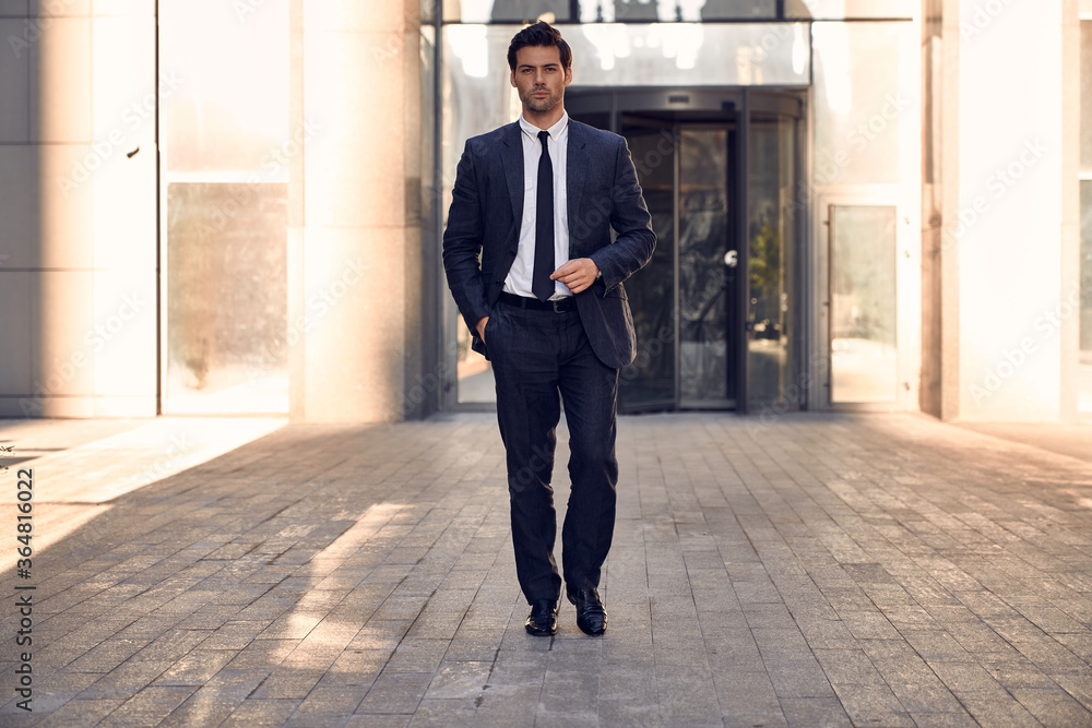 Obraz premium Young and successful. Handsome young businessman adjusting his jacket and looking away while walking outdoors with office building in the background